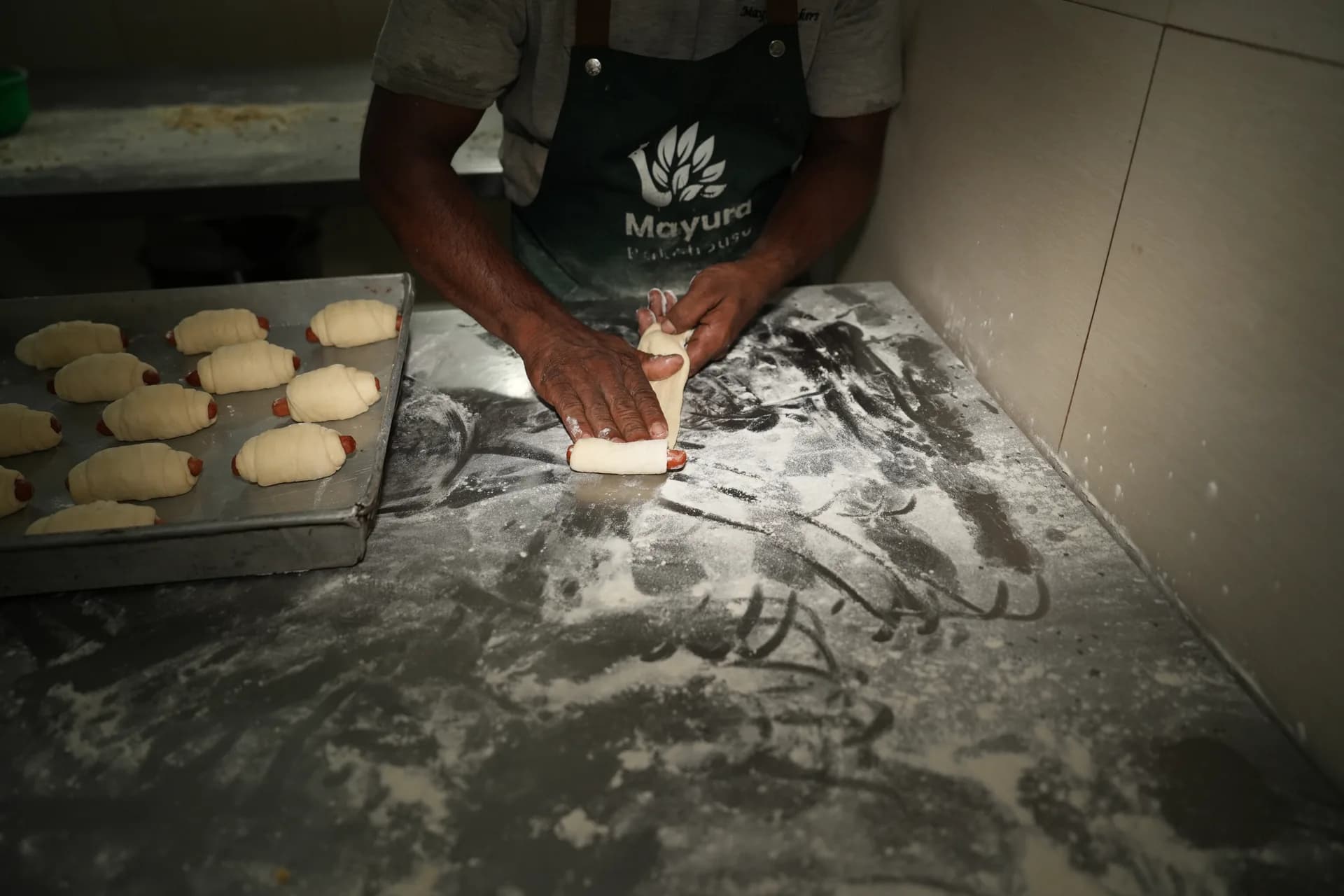 A baker cutting dough on a floured work surface beside a tray of rolls.