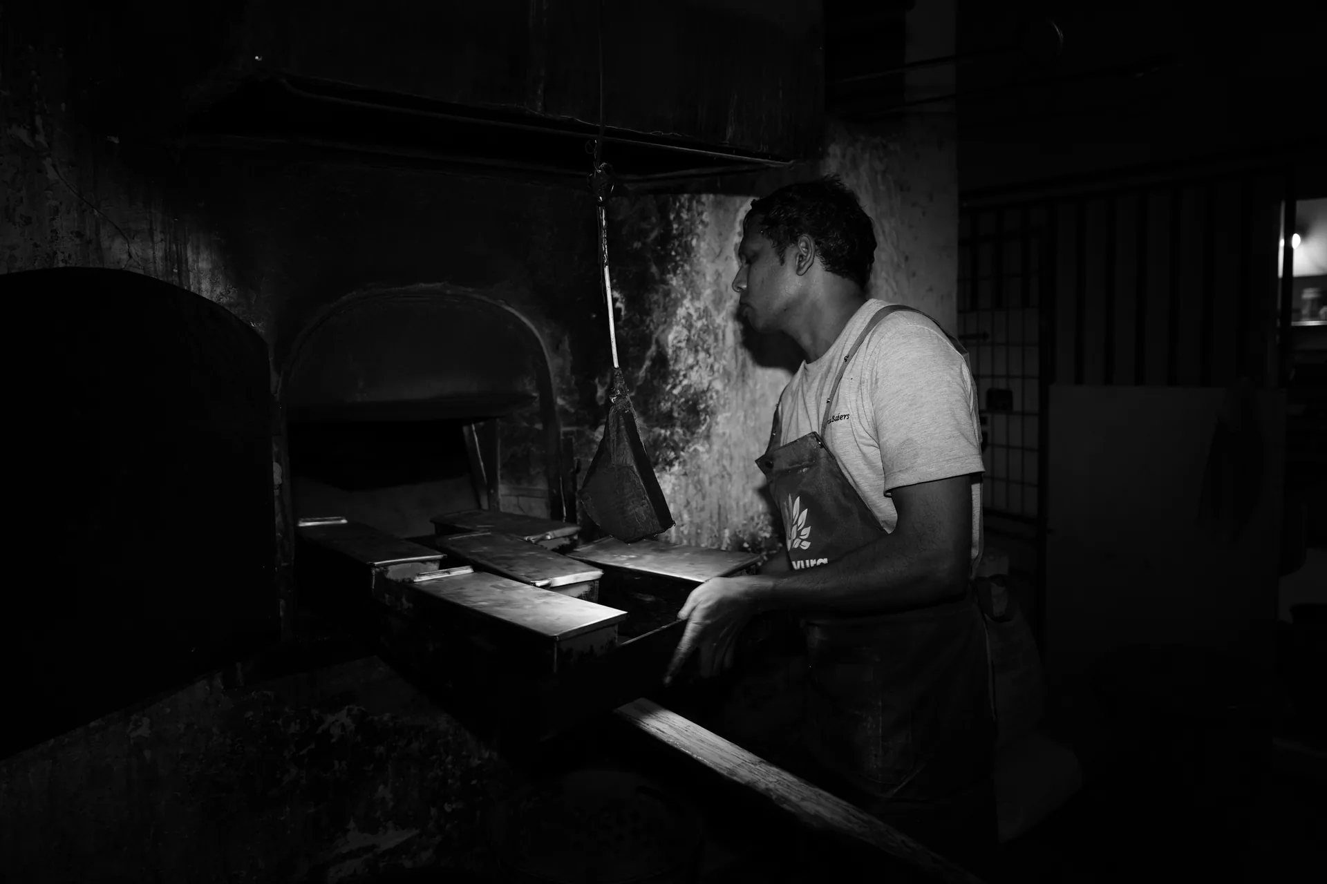 A baker loading trays of bread beside a time-worn brick oven.